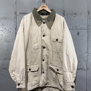 May include: A tan jacket with a green corduroy collar, four front pockets, and black buttons. The jacket is hanging on a wooden hanger against a gray metal background. The jacket appears to be made of a durable, possibly canvas, material.