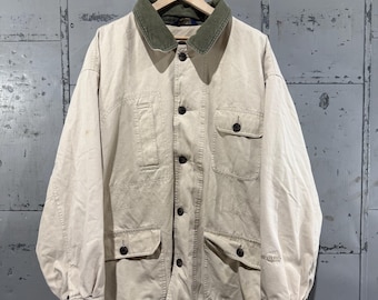 May include: A tan jacket with a green corduroy collar, four front pockets, and black buttons. The jacket is hanging on a wooden hanger against a gray metal background. The jacket appears to be made of a durable, possibly canvas, material.