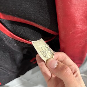 Op de afbeelding: Close-up van een vintage rode en zwarte jas. De jas heeft een rode leren buitenkant en een zwarte wollen binnenkant met rode bies. Een label met de materiaalsamenstelling van de jas is zichtbaar.