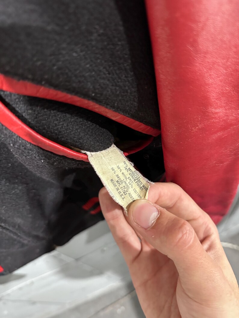 Op de afbeelding: Close-up van een vintage rode en zwarte jas. De jas heeft een rode leren buitenkant en een zwarte wollen binnenkant met rode bies. Een label met de materiaalsamenstelling van de jas is zichtbaar.