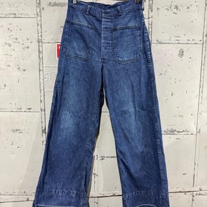 May include: Blue denim pants with large front pockets and a wide leg. The pants are hanging on a wooden hanger against a textured, light-colored background. The hemline features a white accent stitch.