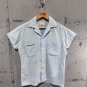 May include: Light blue short-sleeve shirt with two chest pockets and a camp collar. The shirt is buttoned up and hangs on a wooden hanger. The shirt has a textured appearance and is made of a lightweight fabric. The shirt is vintage.