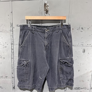 May include: Dark gray cargo shorts with multiple pockets and a button closure. The shorts are hanging on a wooden hanger against a textured metal background. The shorts have a subtle pattern.