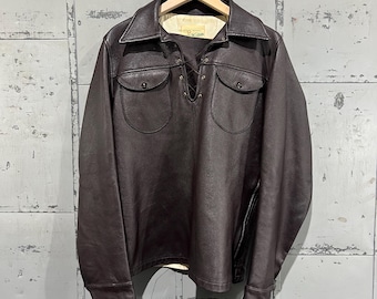 May include: A brown leather shirt with a lace-up front and two flap pockets. The shirt has long sleeves and a collared neckline.