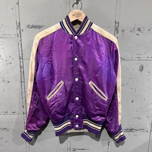 May include: A vintage purple satin bomber jacket with cream-colored sleeves and trim. The jacket features white buttons, two front pockets, and striped cuffs and collar. The jacket is hanging on a metal hook.
