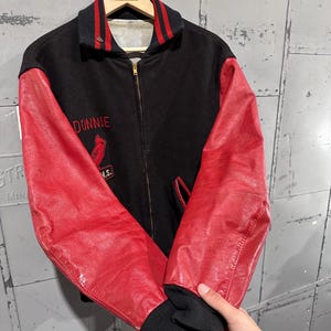 Op de afbeelding: Zwarte en rode vintage varsity-jas met de naam "Donnie" geborduurd op de borst. De jas heeft rode leren mouwen, een zwarte body en een rood-zwarte gestreepte kraag en manchetten. Een kardinaal-logo is ook geborduurd.