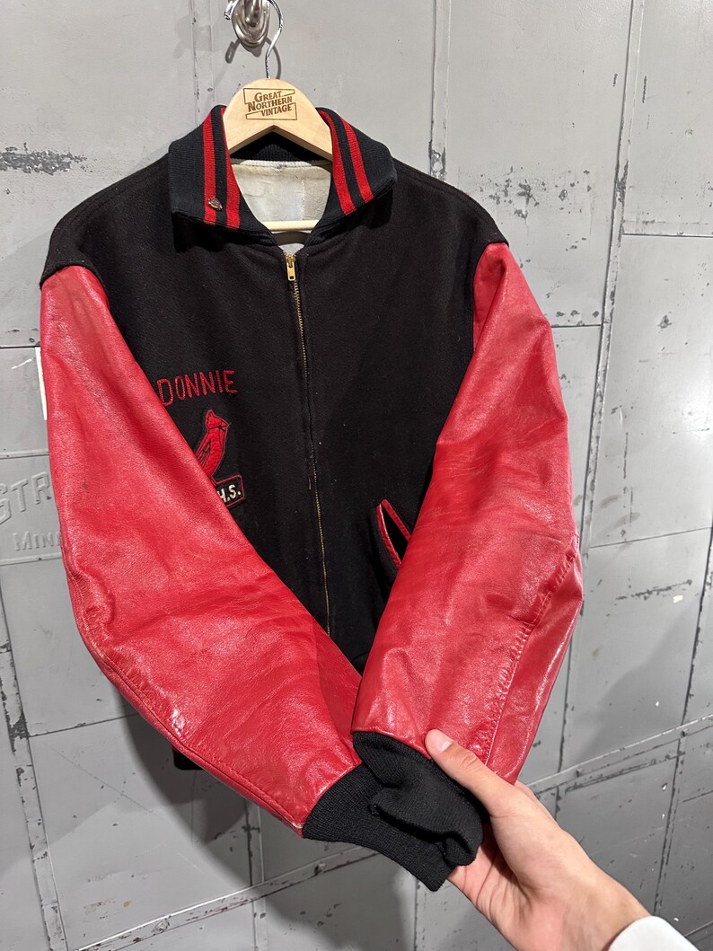 Op de afbeelding: Zwarte en rode vintage varsity-jas met de naam "Donnie" geborduurd op de borst. De jas heeft rode leren mouwen, een zwarte body en een rood-zwarte gestreepte kraag en manchetten. Een kardinaal-logo is ook geborduurd.