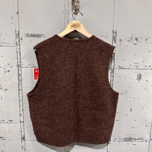 K&ouml;nnte beinhalten: Eine dunkelbraune &auml;rmellose Strickweste mit vertikalem Rippstrickmuster. Das Kleidungsst&uuml;ck hat einen Rundhalsausschnitt und wird auf einem Holzkleiderb&uuml;gel pr&auml;sentiert. Die Textur des Strickmaterials wirkt dick und flauschig und weist durchgehend eine einheitliche Farbe auf.