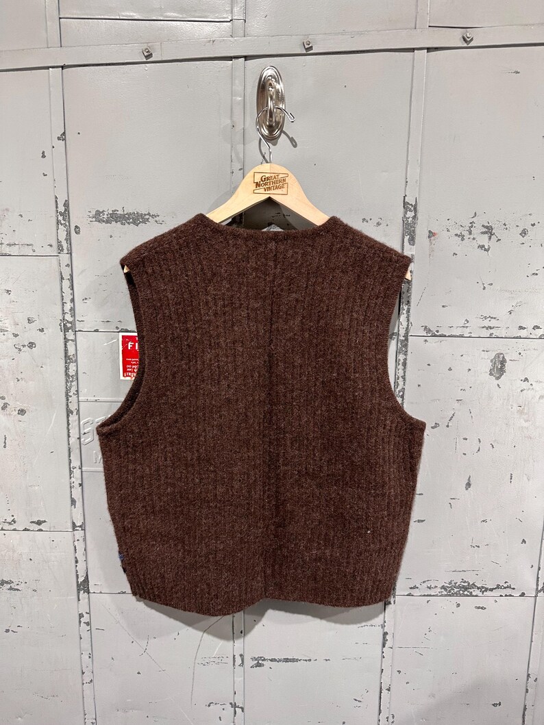 K&ouml;nnte beinhalten: Eine dunkelbraune &auml;rmellose Strickweste mit vertikalem Rippstrickmuster. Das Kleidungsst&uuml;ck hat einen Rundhalsausschnitt und wird auf einem Holzkleiderb&uuml;gel pr&auml;sentiert. Die Textur des Strickmaterials wirkt dick und flauschig und weist durchgehend eine einheitliche Farbe auf.