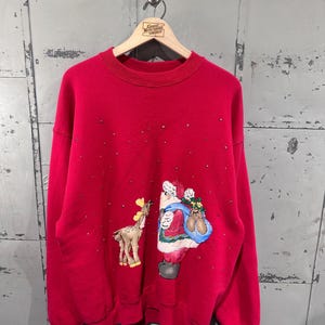 May include: A red crew neck sweatshirt with a festive Christmas design. The graphic features Santa Claus with a sack of gifts and a reindeer. The sweatshirt is adorned with small, scattered embellishments.