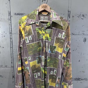 Puede incluir: Una camisa abotonada de manga larga con un estampado temático de la naturaleza. La camisa presenta un patrón repetido de escenas de bosque y texturas de madera en tonos marrones, verdes y amarillos. La camisa está colgada en una percha de madera.