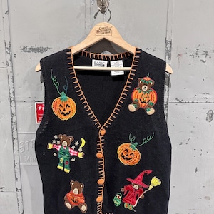 May include: Black knit Halloween vest with orange stitching and button closures. The vest features embroidered pumpkins, bears in costumes, and a witch with a broom. The vest is hanging on a wooden hanger.