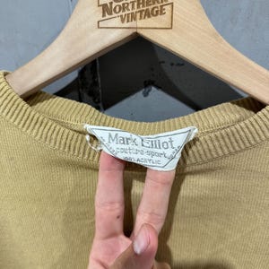 May include: A tan-colored sweater with a ribbed neckline and cuffs, hanging on a wooden hanger. The hanger is branded with "Great Northern Vintage." The sweater's label reads "Mark Elliot couture-sport 100% Acrylic."