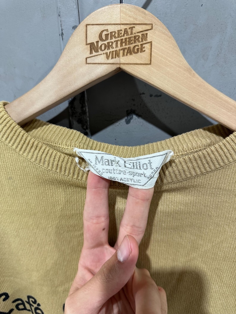 May include: A tan-colored sweater with a ribbed neckline and cuffs, hanging on a wooden hanger. The hanger is branded with "Great Northern Vintage." The sweater's label reads "Mark Elliot couture-sport 100% Acrylic."