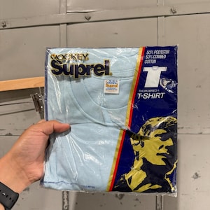 Puede incluir: Una camiseta Jockey Suprel azul claro en su embalaje original. El embalaje es azul y presenta los logotipos de Jockey y Suprel, junto con el texto "50% Poliéster 50% Algodón Peinado" y "Thorobred T-Shirt".