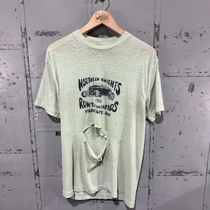 May include: A light green short-sleeve t-shirt with a large hole in the center. The graphic features a black illustration of a hot rod car, the text "NORTHERN KNIGHTS", "1988", "RUN TO THE RAPIDS", and "PARK RAPIDS, MN" printed in black ink.