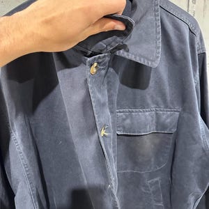 May include: A navy blue jacket with a collared neckline and two front pockets. The jacket has gold-colored buttons and a classic design. The garment is hanging on a wooden hanger.
