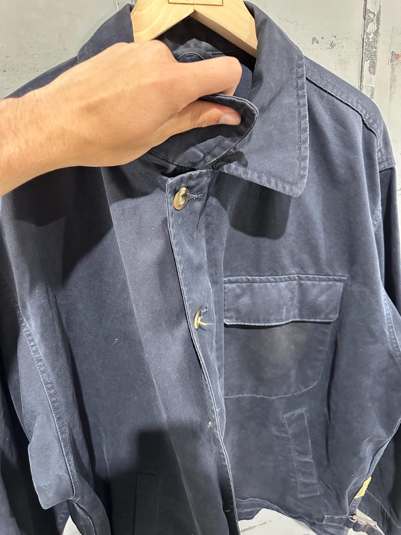 May include: A navy blue jacket with a collared neckline and two front pockets. The jacket has gold-colored buttons and a classic design. The garment is hanging on a wooden hanger.
