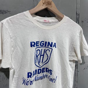 May include: Cream-colored t-shirt with blue text and graphics. The shirt features the words "REGINA", "RHS", "RAIDERS", and "We're Number One!". The shirt is hanging on a wooden hanger with the text "GREAT NORTHERN VINTAGE".