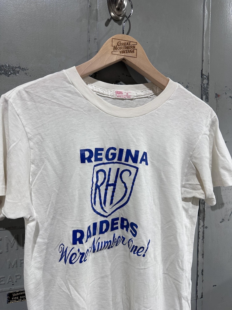 May include: Cream-colored t-shirt with blue text and graphics. The shirt features the words "REGINA", "RHS", "RAIDERS", and "We're Number One!". The shirt is hanging on a wooden hanger with the text "GREAT NORTHERN VINTAGE".