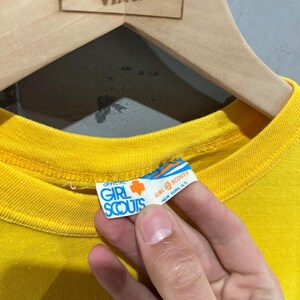 May include: Close-up of a bright yellow t-shirt with the official Girl Scouts tag visible. The tag is white with blue and orange text and a Girl Scout trefoil logo. The shirt is hanging on a wooden hanger.