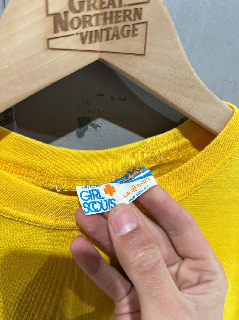 May include: Close-up of a bright yellow t-shirt with the official Girl Scouts tag visible. The tag is white with blue and orange text and a Girl Scout trefoil logo. The shirt is hanging on a wooden hanger.