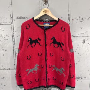 May include: A bright red knit cardigan featuring a repeating pattern of black and grey horses and black horseshoes. The long-sleeved garment has a round neckline, a button-down front closure, and dark trim along the edges of the collar and cuffs.