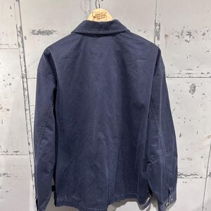 May include: Navy blue work jacket hanging on a wooden hanger. The jacket has a button-down front, long sleeves, and a collar. The jacket is made of a durable fabric and appears to be vintage. The hanger has the text "Great Northern Vintage".