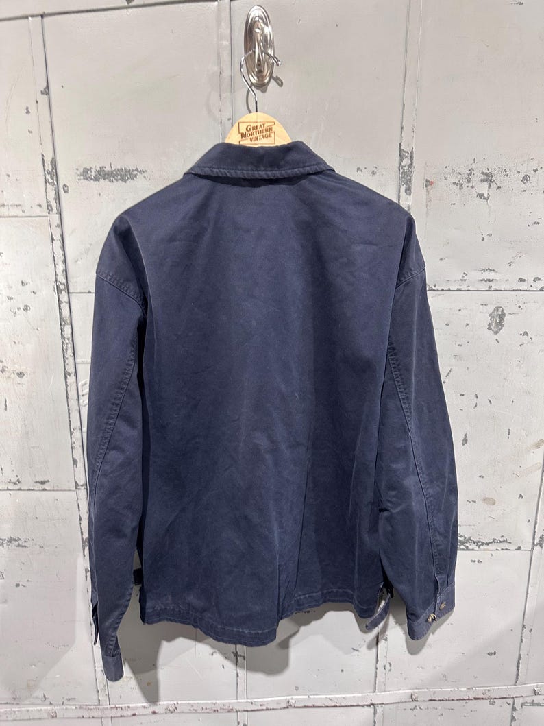 May include: Navy blue work jacket hanging on a wooden hanger. The jacket has a button-down front, long sleeves, and a collar. The jacket is made of a durable fabric and appears to be vintage. The hanger has the text "Great Northern Vintage".