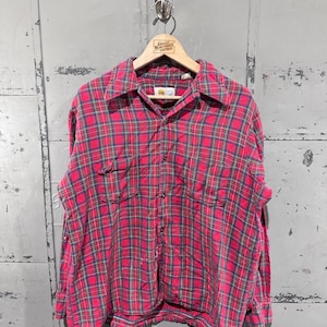 May include: A red and blue plaid button-down shirt with two chest pockets. The shirt is hanging on a wooden hanger with the text "Great Northern Vintage". The shirt is made of a soft, possibly flannel, material.