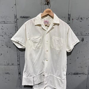 Pode incluir: Camisa creme de manga curta com dois bolsos frontais e fecho de botões. A camisa está pendurada em um cabide de madeira com o texto "Great Northern Vintage". A camisa tem um corte folgado e uma gola clássica.