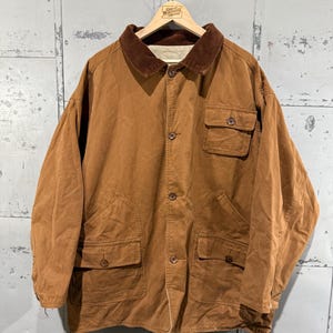 May include: A brown corduroy jacket with a brown corduroy collar and four front pockets. The jacket is buttoned up and hanging on a wooden hanger. The hanger has a small sign that reads "Great Northern Vintage".