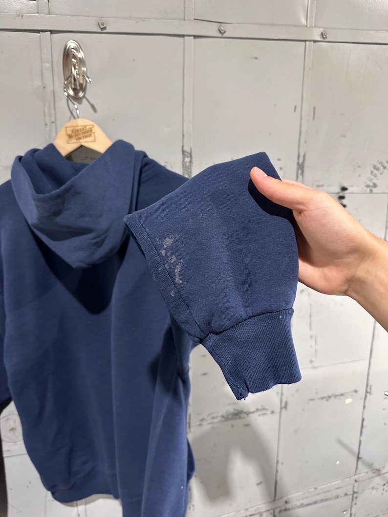 May include: Navy blue hoodie with a drawstring hood and ribbed cuffs. The sleeve shows discoloration and a small tear. The hoodie is hanging on a wooden hanger.