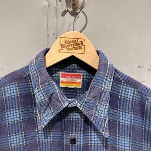 Pu&ograve; includere: Una camicia a maniche lunghe con bottoni caratterizzata da un motivo a quadri blu e azzurro con sottili linee gialle. La camicia presenta un colletto a punta e un'etichetta rettangolare all'interno del collo con la scritta FLANNELCRAFT 100% COTTON.
