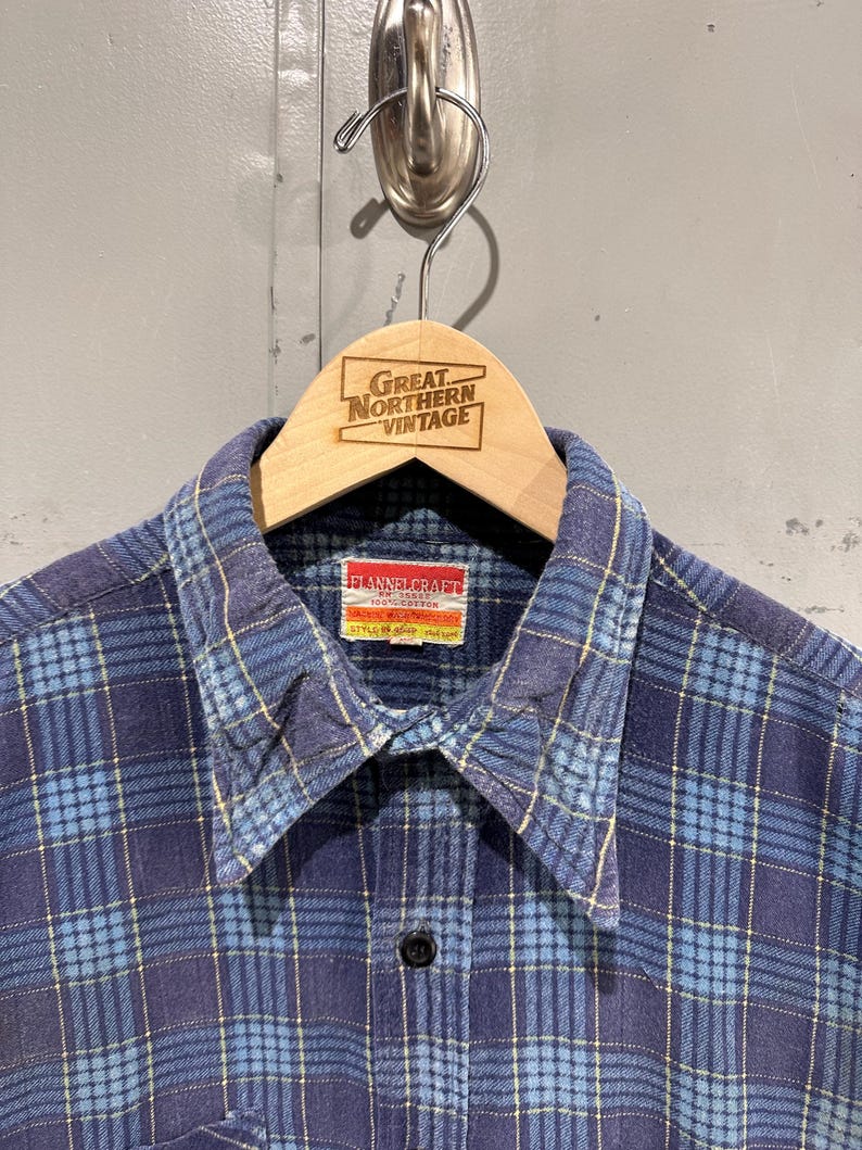 Pu&ograve; includere: Una camicia a maniche lunghe con bottoni caratterizzata da un motivo a quadri blu e azzurro con sottili linee gialle. La camicia presenta un colletto a punta e un'etichetta rettangolare all'interno del collo con la scritta FLANNELCRAFT 100% COTTON.
