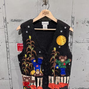 May include: Black knit vest with a fall harvest theme. Features embroidered scarecrows, pumpkins, corn stalks, and a full moon. The vest has a button-up front and is decorated with star-shaped accents.