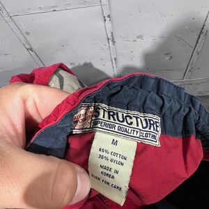 May include: A red and navy blue clothing label with the text "STRUCTURE SUPERIOR QUALITY CLOTHING" and "M 65% COTTON 35% NYLON MADE IN KOREA QUER FOR CARE".