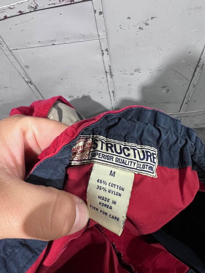 May include: A red and navy blue clothing label with the text "STRUCTURE SUPERIOR QUALITY CLOTHING" and "M 65% COTTON 35% NYLON MADE IN KOREA QUER FOR CARE".