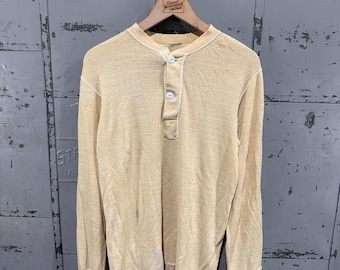 May include: A pale yellow long-sleeve henley shirt featuring a three-button placket with white buttons, a round neckline, and white contrast stitching along the seams. The fabric has a textured appearance with subtle signs of wear and discoloration.
