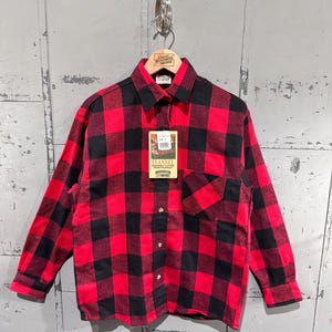 May include: A red and black buffalo plaid flannel shirt with a button-down front and a chest pocket. The shirt is hanging on a metal hook against a weathered, gray background. A tag reads "Flannel, Heavyweight".