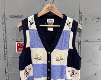 May include: A vintage, button-down sweater vest with a patchwork design. The vest features navy blue, cream, and light blue panels, with embroidered floral details in yellow and pink. The vest is sleeveless and hangs on a wooden hanger.