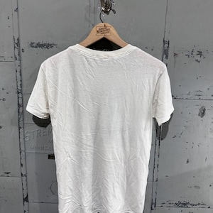May include: A white, short-sleeved t-shirt hanging on a wooden hanger. The shirt is plain and appears to be made of a soft, lightweight fabric. The hanger has a logo that reads "Great Northern Vintage". The background is a weathered, gray metal surface.