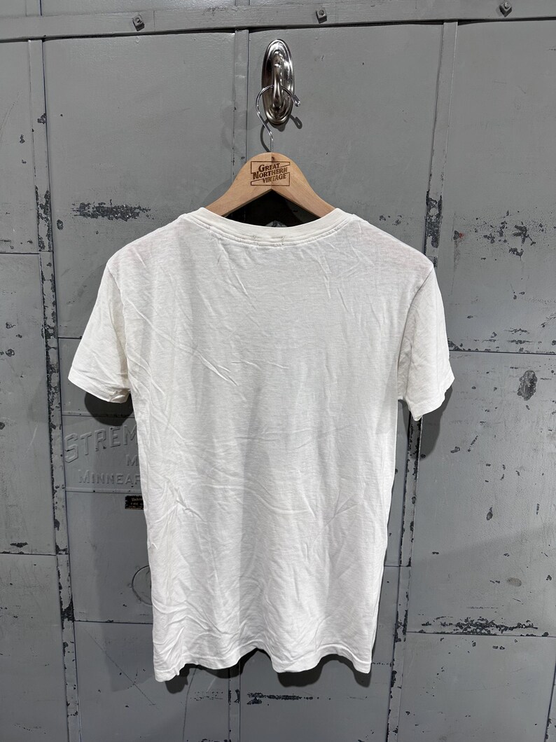 May include: A white, short-sleeved t-shirt hanging on a wooden hanger. The shirt is plain and appears to be made of a soft, lightweight fabric. The hanger has a logo that reads "Great Northern Vintage". The background is a weathered, gray metal surface.