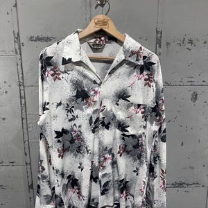 May include: A vintage button-down shirt with a floral pattern. The shirt is white with a design of black, gray, and pink leaves. The shirt has long sleeves and a single pocket on the chest. The shirt is hanging on a wooden hanger.