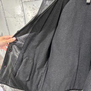 May include: A black leather jacket featuring a dark gray interior lining. The jacket has a notched collar, a button-front closure, and a visible interior pocket on the left side. A black fabric care label is attached near the collar.