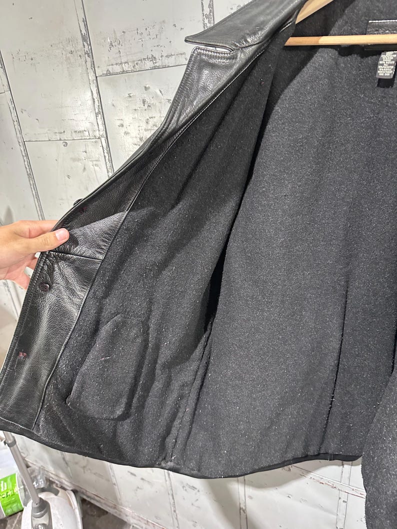 May include: A black leather jacket featuring a dark gray interior lining. The jacket has a notched collar, a button-front closure, and a visible interior pocket on the left side. A black fabric care label is attached near the collar.