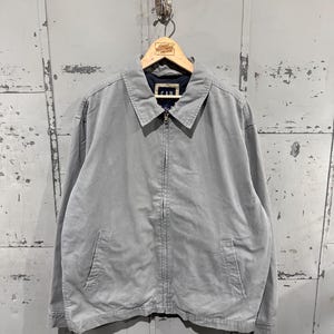 May include: A light gray zip-up jacket with a collar and two front pockets. The jacket is hanging on a wooden hanger with the GAP logo visible. The jacket is made of a soft, durable material.
