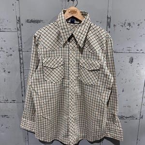 May include: A long-sleeved, button-down shirt with a plaid pattern in shades of beige, green, and black. The shirt features two chest pockets and a pointed collar. The shirt is hanging on a wooden hanger.