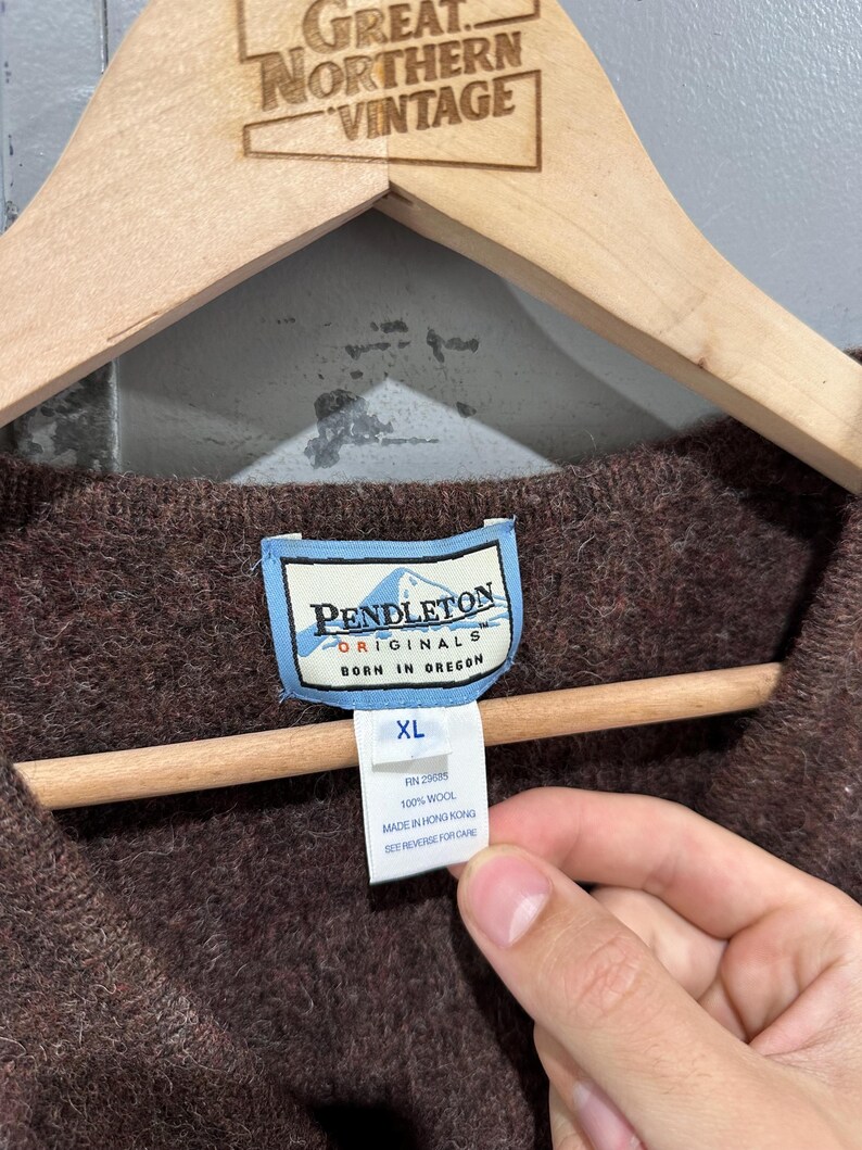 K&ouml;nnte beinhalten: Ein brauner Wollpullover mit einem Pendleton Originals Etikett mit dem Text Pendleton Originals Born in Oregon. Ein wei&szlig;es Zusatzetikett zeigt die Gr&ouml;&szlig;e XL, RN 29685, 100% Wool, Made in Hong Kong und den Hinweis See Reverse for Care.