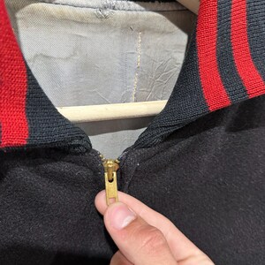 Op de afbeelding: Close-up van een zwarte jas met een rood-zwarte gestreepte kraag. De jas heeft een gouden rits en een grijze voering. De jas hangt aan een houten kleerhanger.